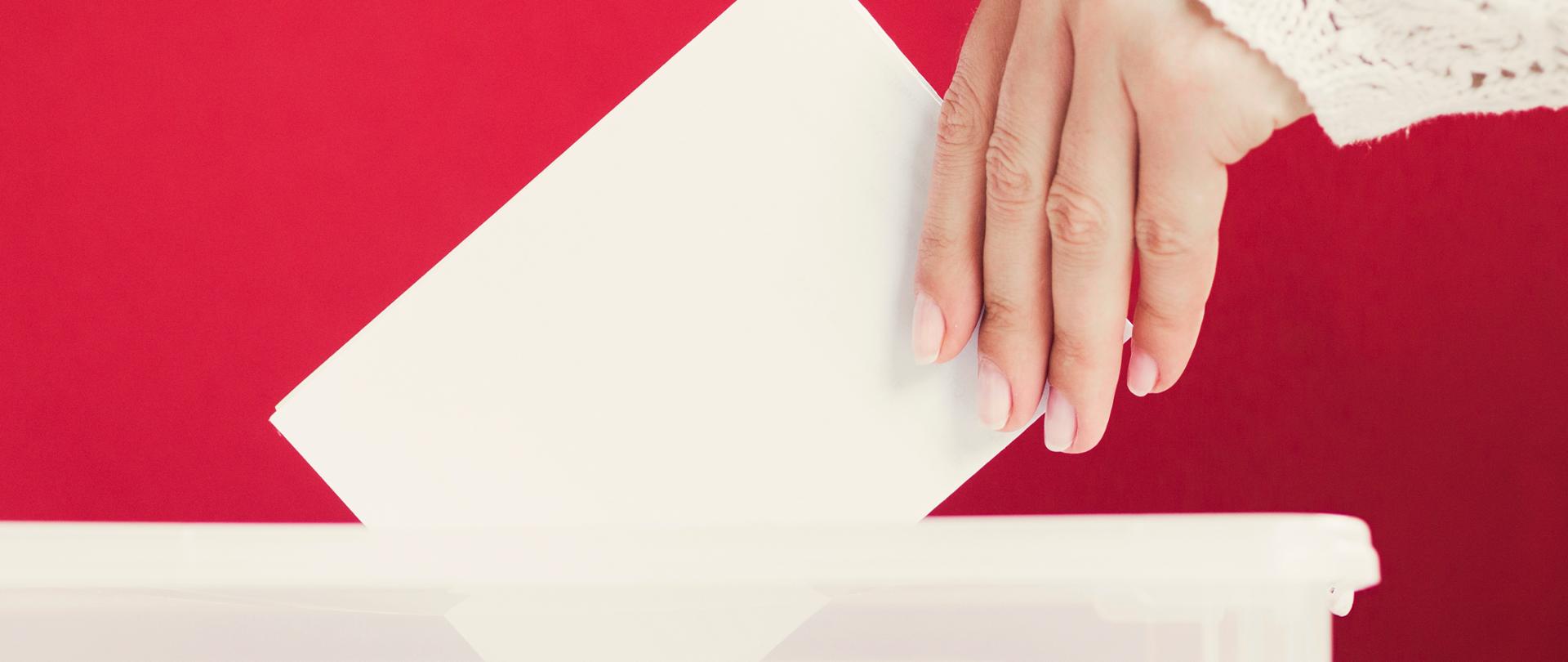 Zdjęcie przedstawia kobiecą dłoń, która wrzuca białą kartkę do urny.