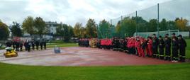 Na fotografii rzut ogólny na stadion. Na tartanowej nawierzchni ustawione w dwuszeregu wszystkie drużyny biorące udział w zawodach. Na przeciw zawodników stoją organizatorzy i sędziowie. Po środku widać podium oraz stół z nagrodami. W tle widoczny samochód strażacki.
