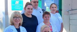 Zdjęcie przedstawia 6 osób, na schodach przed budynkiem Centrum Usług Społecznych Gminy Leżajsk, na co wskazuje logo na na drugim planie w lewym górnym rogu, gdzie na tabliczce o czerwonym tle znajduje się białego koloru napis. obok znajdują się zielonego koloru drzwi do budynku z szybą na środku. 4osoby na schodach to kobiety, które mają założone białe fartuchy z krótkim rękawem i w rękach trzymają granatowe torby, dwie pozostałe osoby to kobieta i mężczyzna ubrani w koszulki - kobieta ma czarny t-shirt, a mężczyzna biały. 