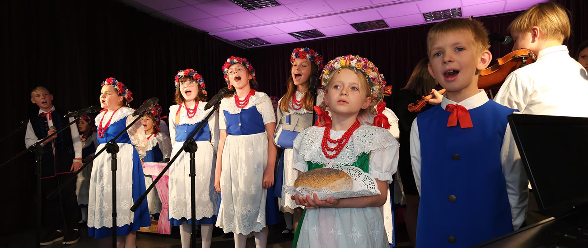 Grupa dzieci w biało niebieskich strojach śpiewająca na scenie
