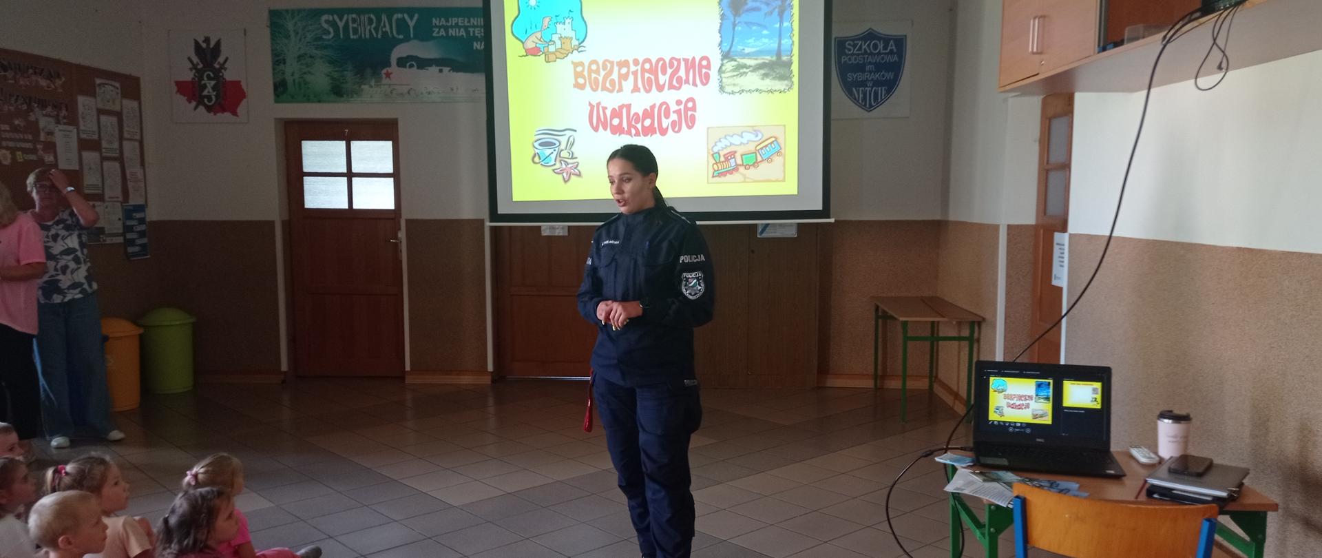 Na środku stoi policjantka ubrana w służbowy mundur. Za nią, na rzutniku wyświetlony jest napis : BEZPIECZNE WAKACJE.