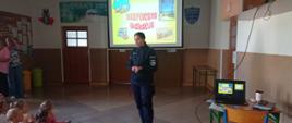 Na środku stoi policjantka ubrana w służbowy mundur. Za nią, na rzutniku wyświetlony jest napis : BEZPIECZNE WAKACJE.