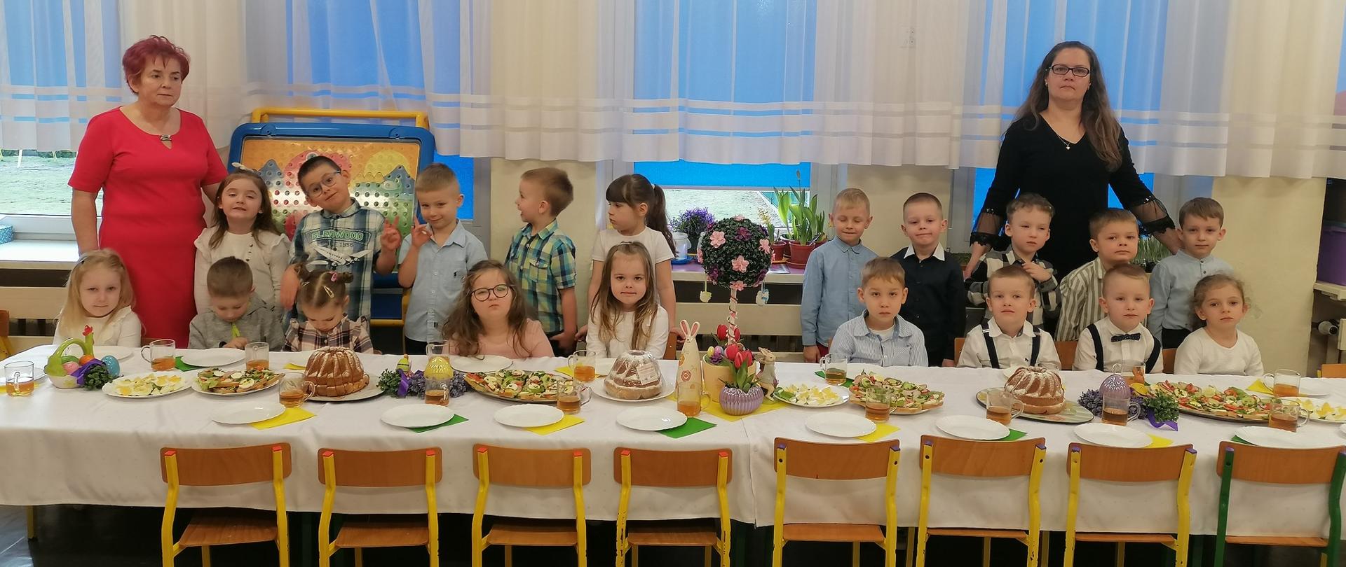 grupa dzieci i 2 nauczycieli stoją za stołem z potrawami wielkanocnymi
