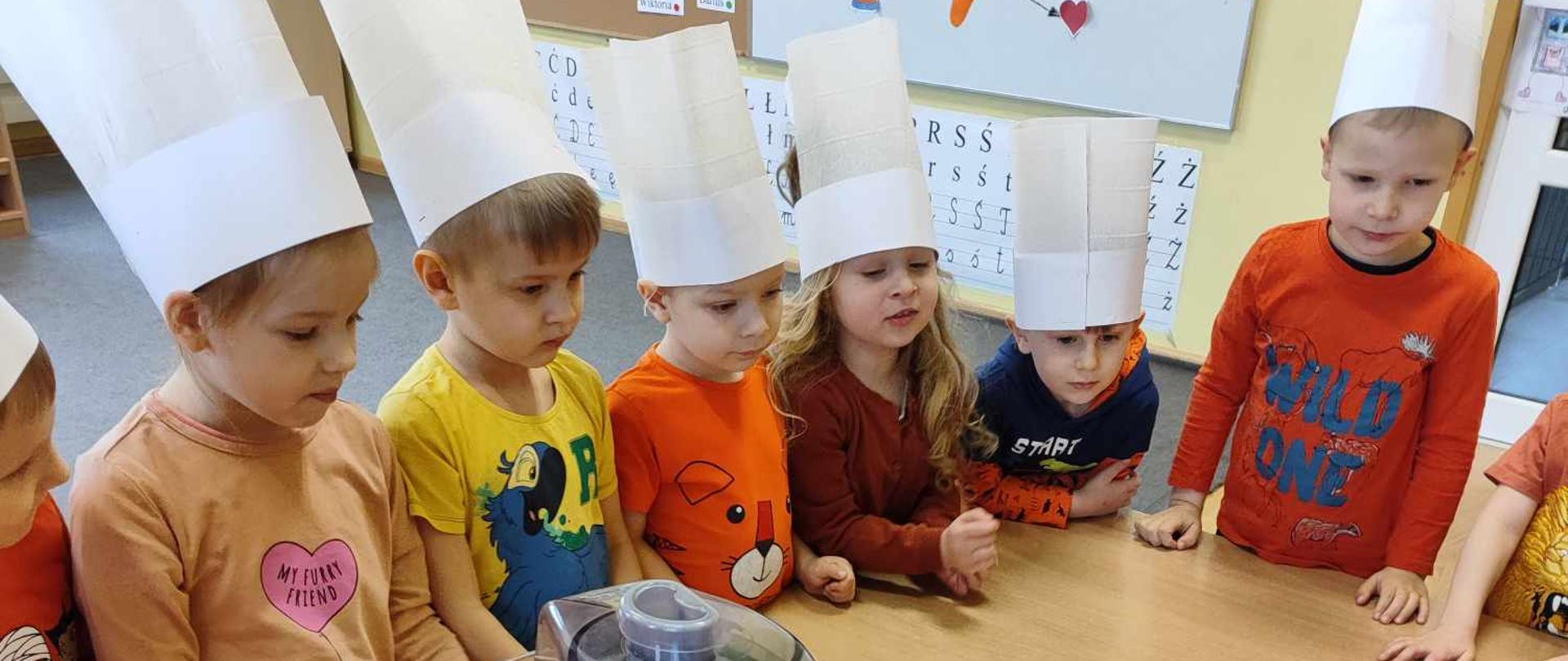 Dzieci ubrane na pomarańczowo w czapkach kucharskich stoją wokół stolika, na którym stoi sokowirówka.