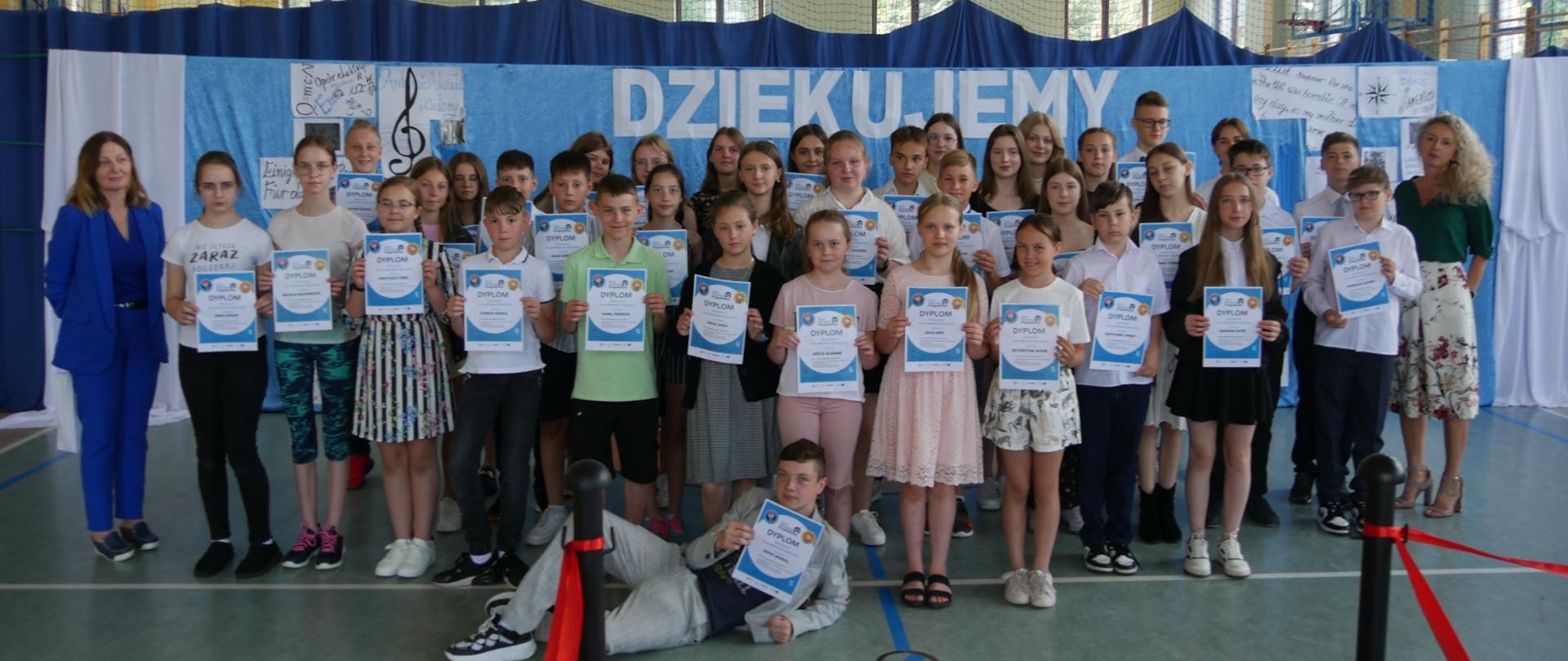 Na hali sportowej ustawieni są obok siebie uczniowie klas I- VIII, którzy zostali wyróżnieni podczas apelu podsumowującego sukcesy uczniów . Każdy z wyróżnionych uczniów trzyma dłoni w dyplom, obok uczniów z prawej strony znajduje się Pani Dyrektor, a z lewej strony Pani Katarzyna Klinowska Banaś.