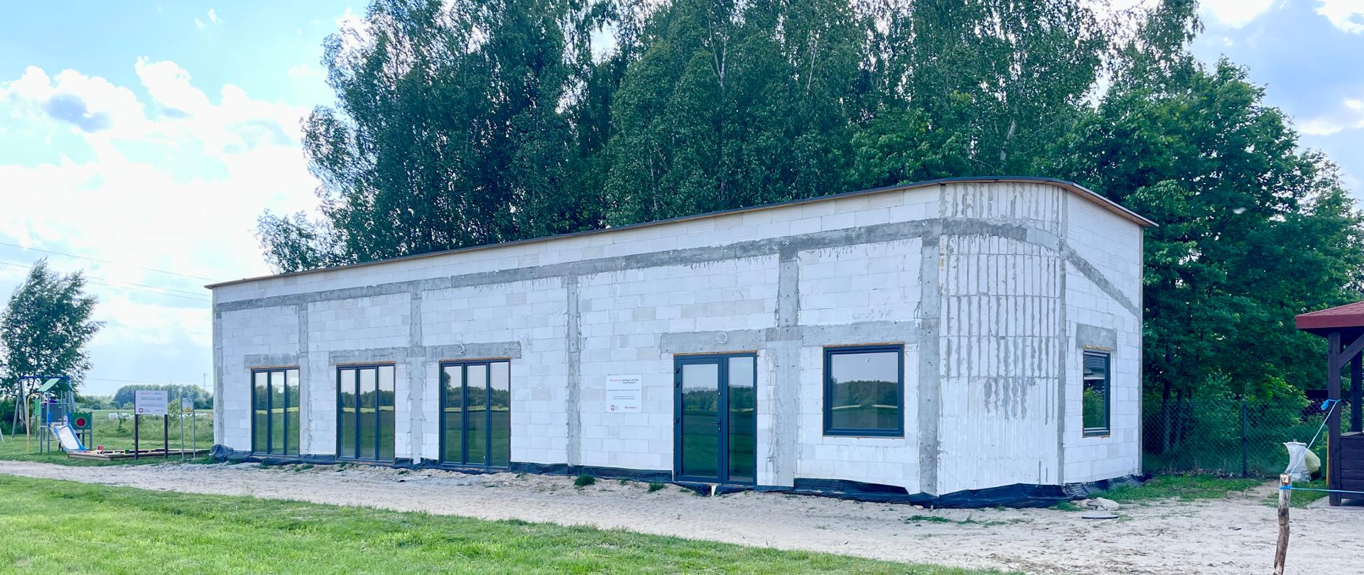 Parterowy budynek w stanie surowym zamkniętym za którym rosną wysokie drzewa w budynku znajduje się 3 duże okna tarasowe. Obok świetlicy jest plac zabaw 