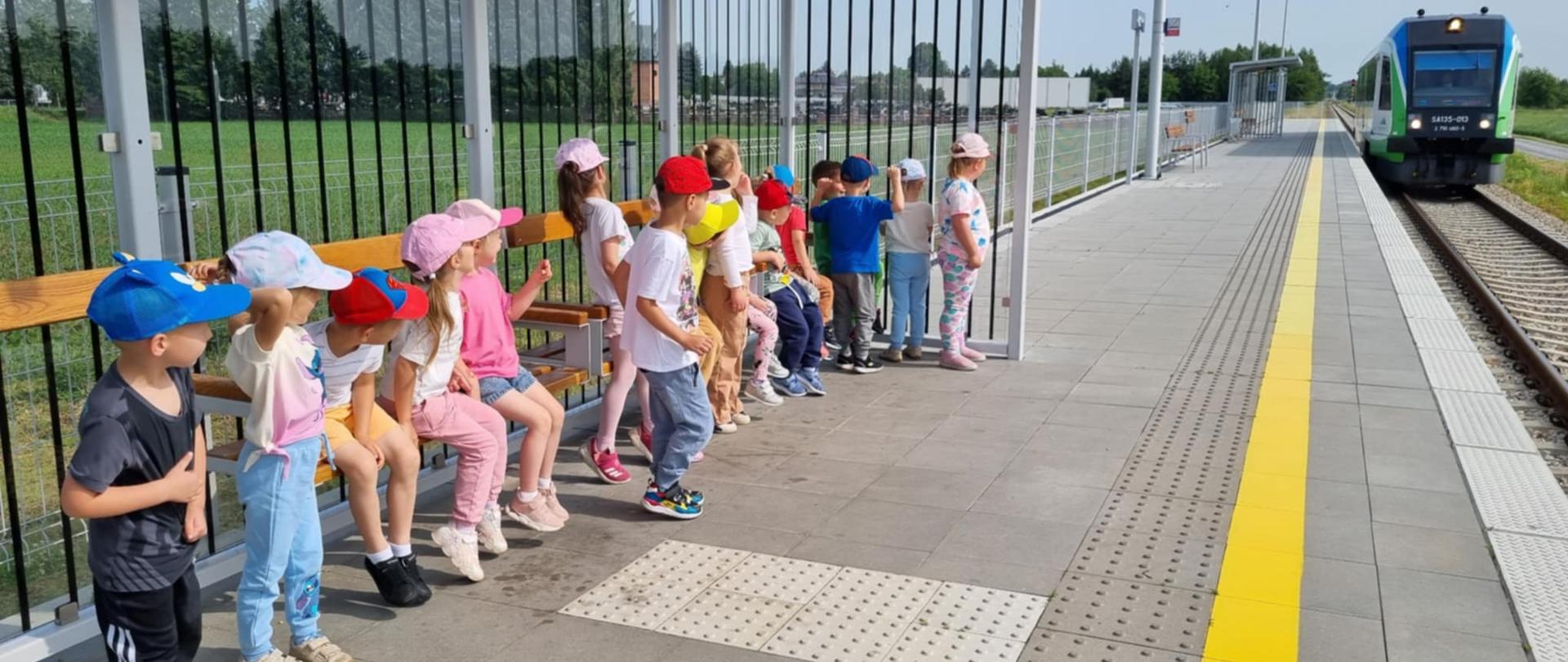 Grupa dzieci oczekuje na pociąg na przystanku kolejowym. W oddali widać nadjeżdżający pociąg.