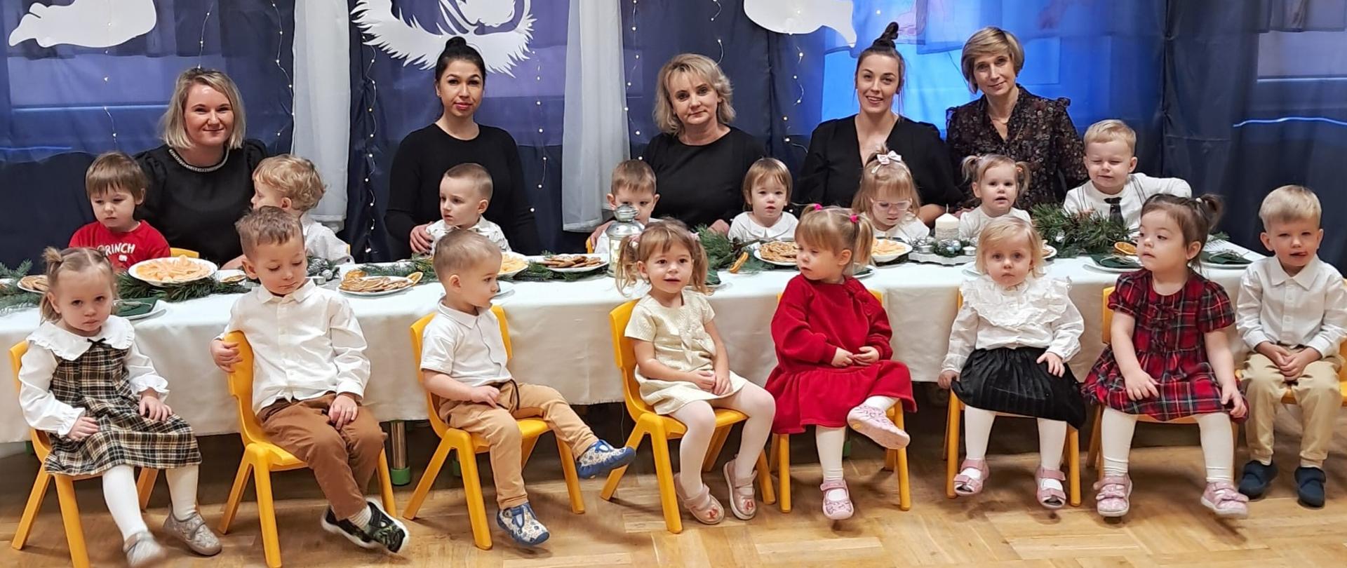 Na zdjęciu widać Panią Dyrektor, ciocie oraz dzieci z grupy Tygryski. Wszyscy są odświętnie ubrani, siedzą dookoła wigilijnego stołu. W tle dekoracja z napisem Radość, Miłość, Pokój...