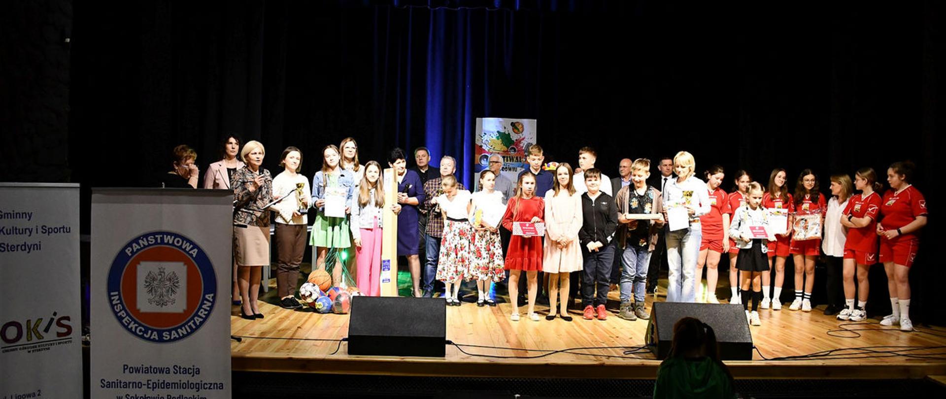Na zdjęciu na oświetlonej scenie z drewnianą podłogą widzimy uczestników konkursu.