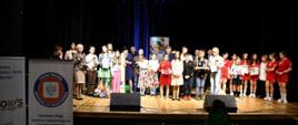 Na zdjęciu na oświetlonej scenie z drewnianą podłogą widzimy uczestników konkursu.