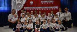 Na zdjęciu widać dzieci oraz ciocie z grupy Tygryski. Wszyscy są ubrani w eleganckie ubrania. W tle dekoracja biało-czerwony materiał imitujący flagę Polski, ilustracja Godła oraz napis Listopada Dzień Niepodległości.