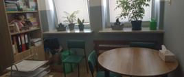 Gabinet pedagoga z jasnymi panelami na podłodze i popielatymi ścianami, po lewej stronie szafka z segregatorami i pomocami dydaktycznymi, po prawej stronie okrągły stolik z zielonymi krzesłami, na wprost okno zasłonięte żaluzjami, na parapecie kwiaty w doniczkach