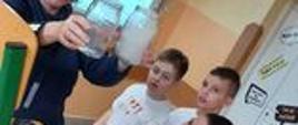 Podczas zajęć - nauczyciel trzyma nad głowami uczniów dwa słoiki - jeden wypełniony cieczą