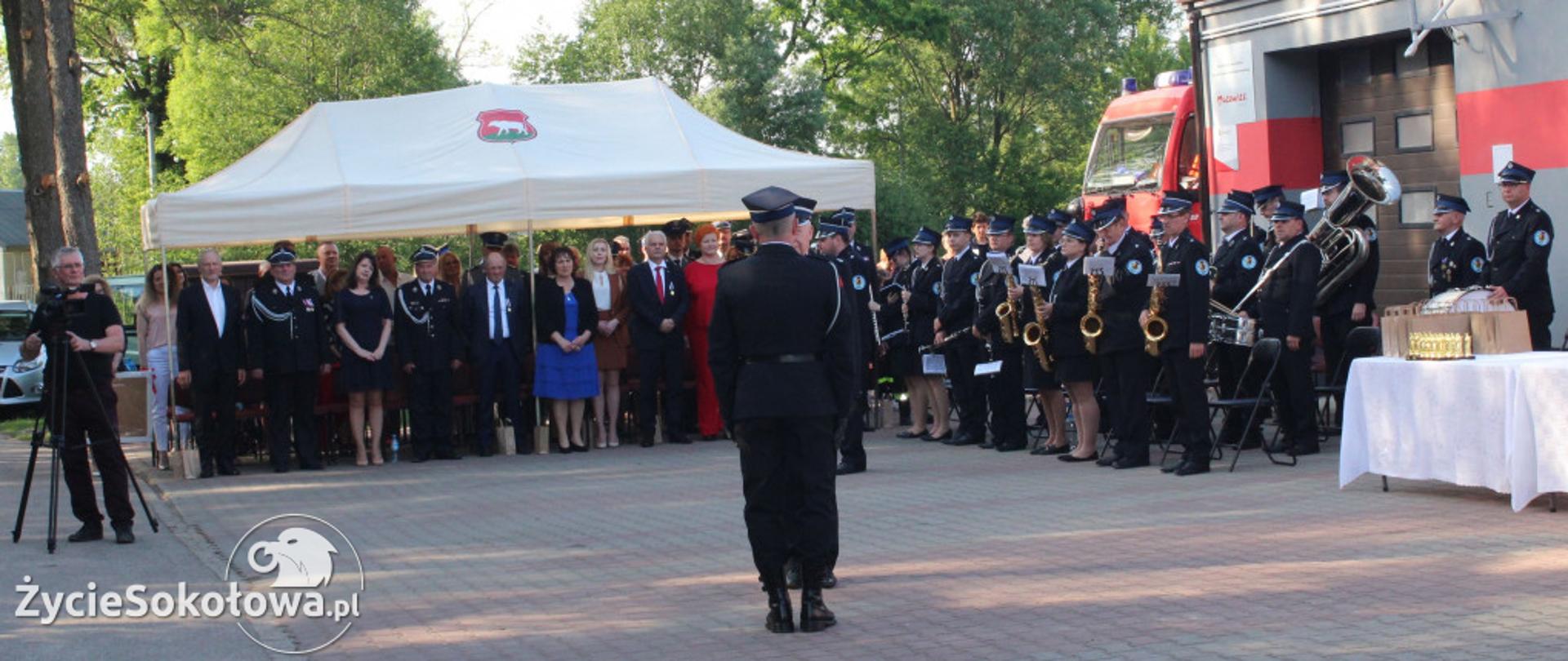 Uroczysty apel na placu przed jednostką OSP w Chruszczewce. Widoczny Moment składania raportu dowódcy. W tle namiot z gośćmi oraz orkiestra dęta OSP Chruszczewka. Po prawej stronie widoczna jednostka oraz fragment strażackiego wozu bojowego.