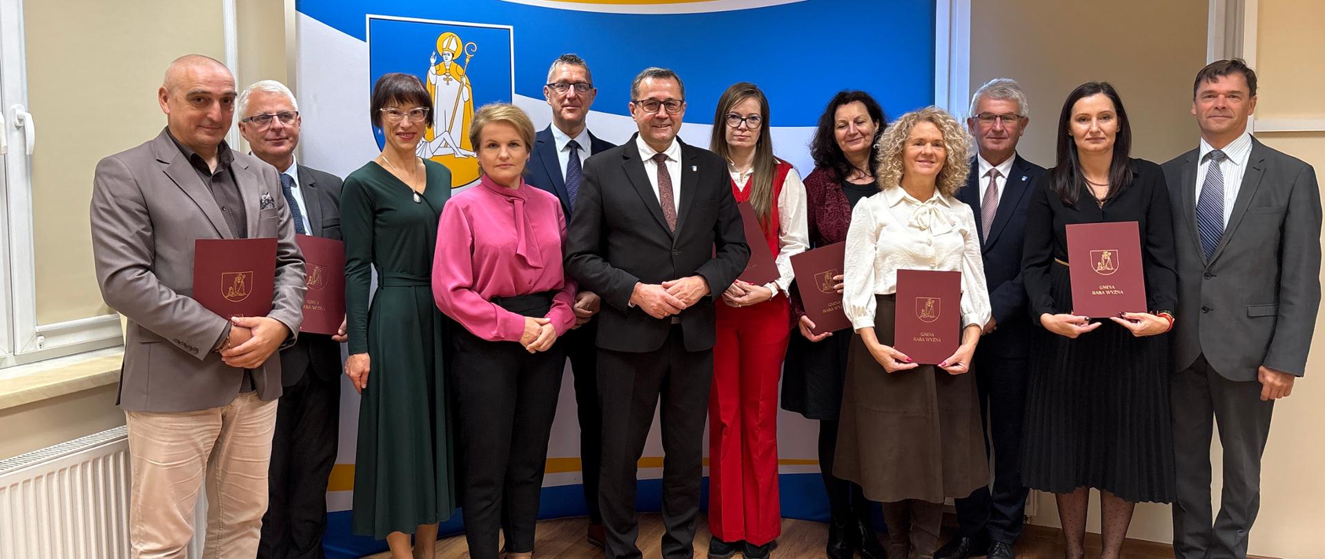 Większa grupa osób w eleganckich ubraniach pozuje do wspólnego zdjęcia. Za nimi żółto niebieska ścianka reklamowa.