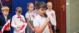 Uczeń klasy 3 odświętnie ubrany recytuje do mikrofonu na akademii, obok stoją inne odświętnie ubrane dzieci