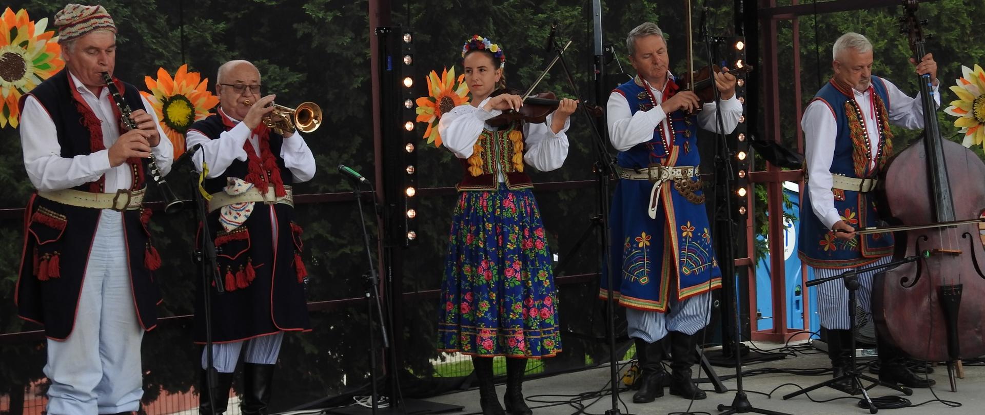 Zdjęcie przedstawia młodą kobietę i 4 mężczyzn w starszym wieku stojących na scenie z różnymi instrumentami. Osoby te ubrane są w tradycyjne stroje ludowe.