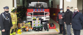Zdjęcie przedstawiające na tle otwartego garażu z widocznym samochodem bojowym i sprzętem ,władze urzędu przekazujący dokumenty zakupu sprzętu strażackiego dla strażaków z Bielanki.