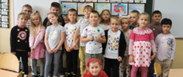Grupa dzieci stoi a za nimi tablica na ścianie z kolorowymi obrazkami