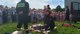 Strażacy wykonują pokaz resuscytacji na trawie, wokół zgromadzone dzieci
