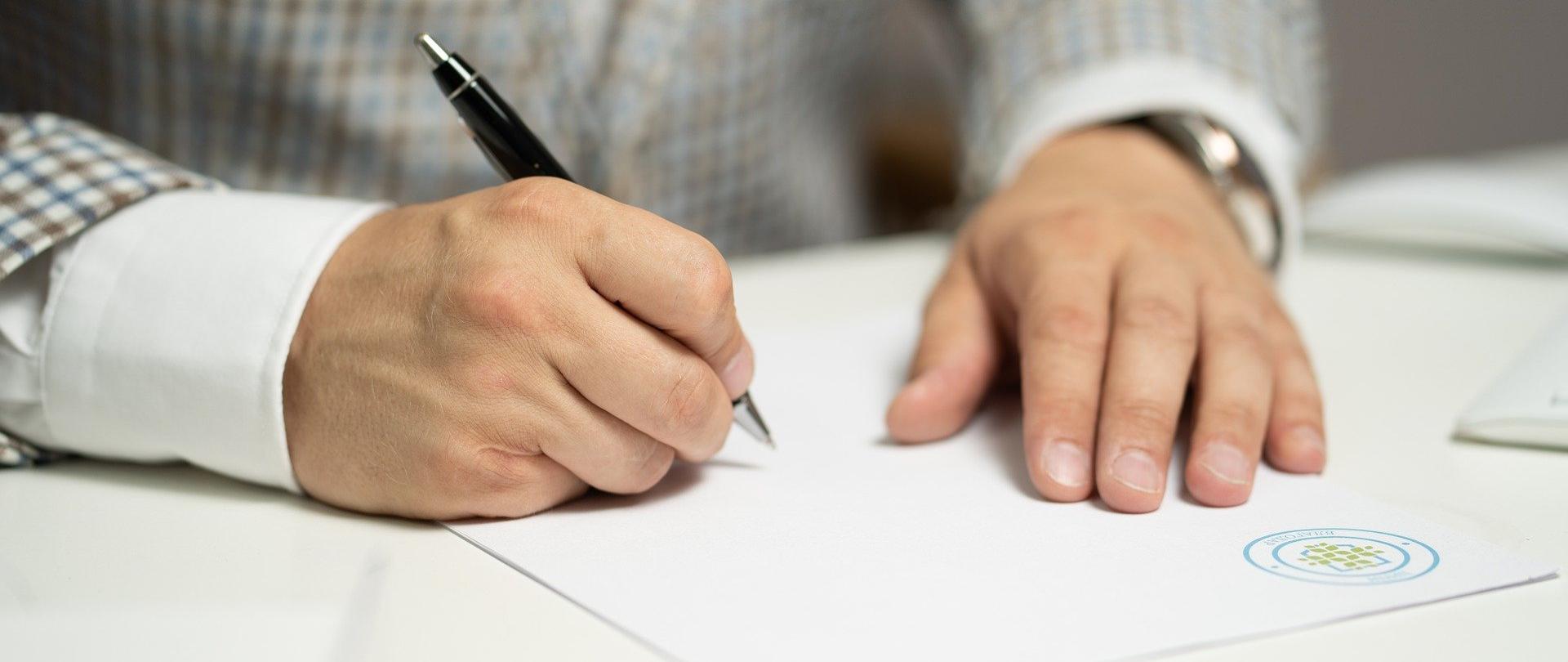 Zdjęcie przedstawia jedną osobę siedzącą przy biurku z długopisem w ręce i piszącą na kartce wewnątrz budynku. Osoba jest przedstawiona w taki sposób, że widoczny jest jedynie jej tułów i dłonie.