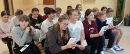 Uczniowie siedzą na ławkach w trzech rzędach, trzymają w ręku kartki z portretem Jana Kochanowskiego i tekstem jego fraszki "Na zdrowie"
