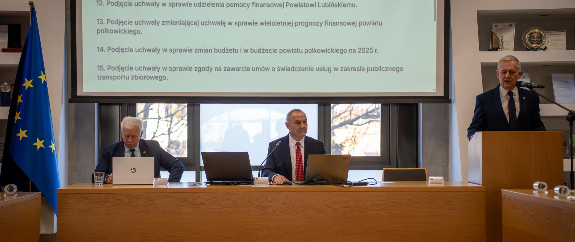 Na zdjęciu widać trzy osoby, dwie siedzą przy stole, jedna przemawia na mównicy