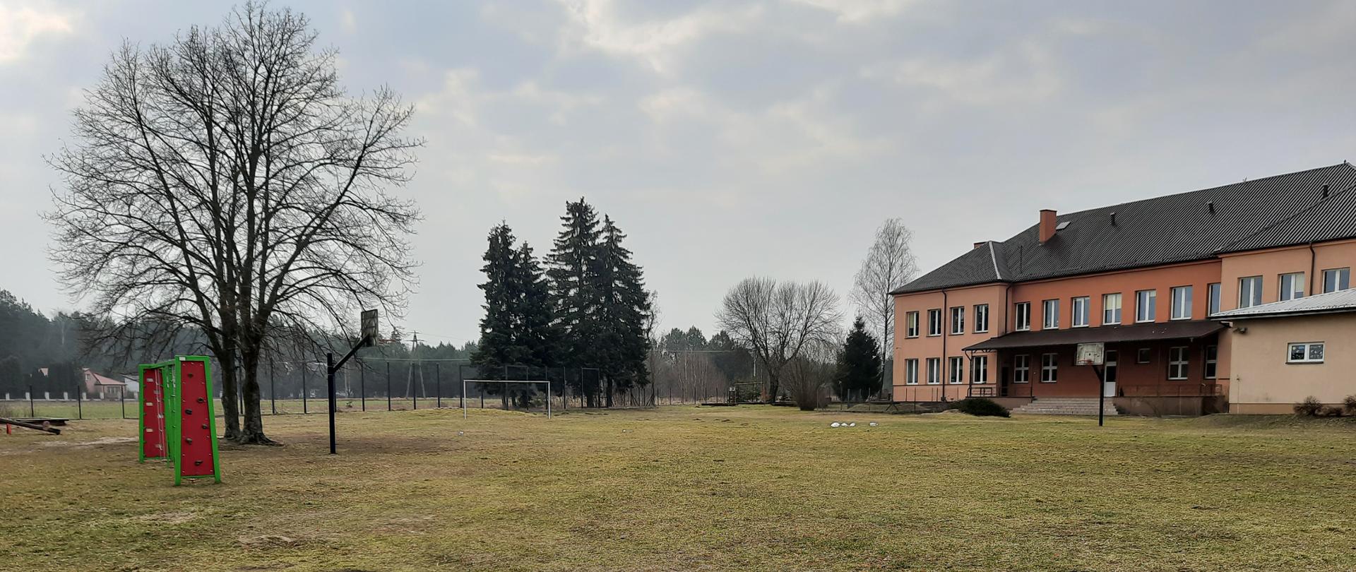 Naturalna nawierzchnia boiska, w oddali bramka, ogrodzenie, las. Po prawej stronie znajduje się budynek szkoły z pomarańczową elewacją.