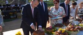 Na zdjęciu podczas plenerowej uroczystości kilka osób prezentuje i kroi duże, ozdobnie wypieczone bochny chleba. Na pierwszym planie znajduje się długi stół zastawiony licznymi bochnami udekorowanymi kwiatami, zielenią i wstążkami. W centrum mężczyzna w garniturze kroi jeden z chlebów, a obok stoją inni uczestnicy wydarzenia. W tle widać scenę z muzykami, zabudowania oraz ogrodzony teren rekreacyjny.