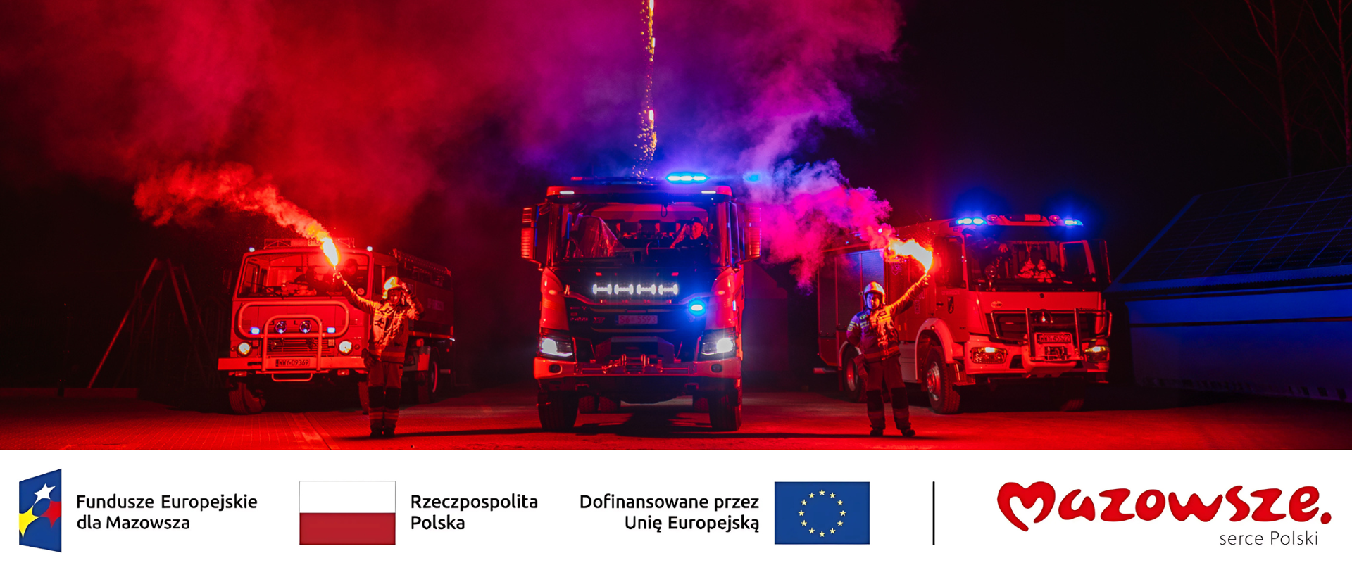 Parking, trzy wozy strażackie, przy nich stoi dwóch strażaków z flarami , w niebo wystrzelone są sztuczne ognie. Jest dużo czerwonego światła i dymu. Poniżej logotypy Funduszy Europejskich dla Mazowsza, Flagi Polski i Unii Europejskiej oraz Mazowsze serce Polski.