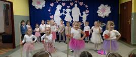 Ubrane w białe bluzeczki i kolorowe spódniczki dziewczynki z przedszkola tańczą taniec z mopami, za nimi stoją chłopcy, w tle na granatowej zasłonie przypięte wycięte z białego papieru postaci babci i dziadka z wnuczką i wnukiem w ramce utworzonej z różowych serduszek, nad nimi napis "Babciu, dziadku, kochamy Was"