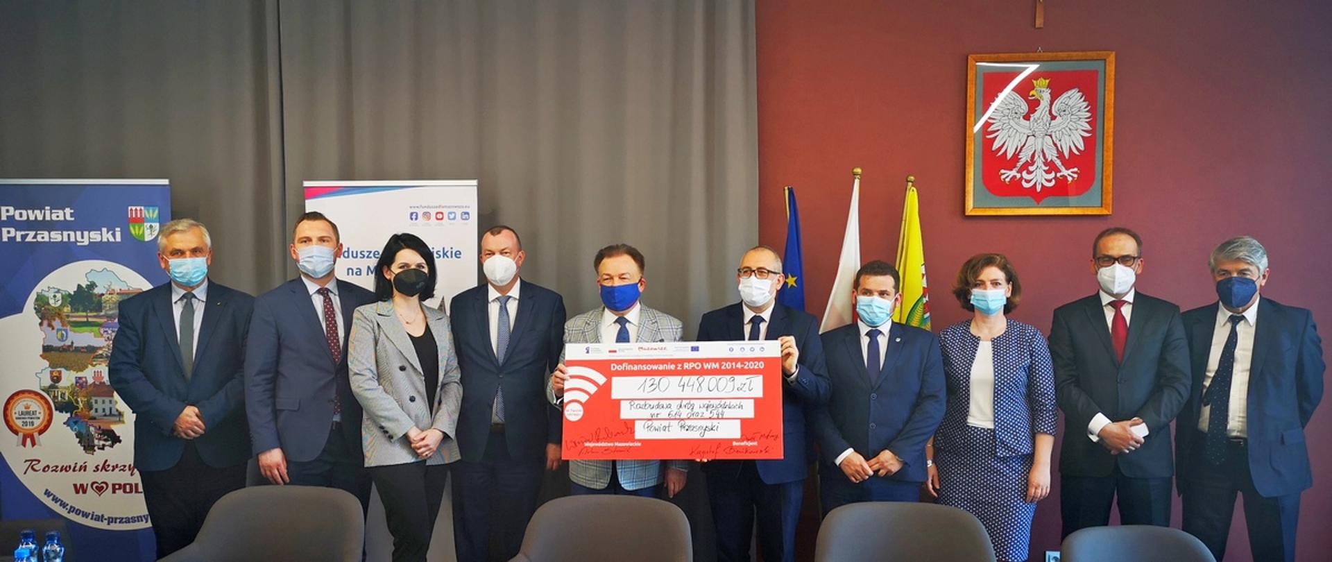 Zdjęcie wykonano w sali konferencyjnej Centrum Aktywizacji Biznesu w przasnyskim starostwie. Na fotografii uwieczniono Marszałka Województwa Mazowieckiego Adama Struzika i Starostę Przasnyskiego Krzysztofa Bieńkowskiego trzymających symboliczny czek. Po prawej i lewej stronie stoją pracownicy starostwa oraz samorządowcy z terenu powiatu przasnyskiego.