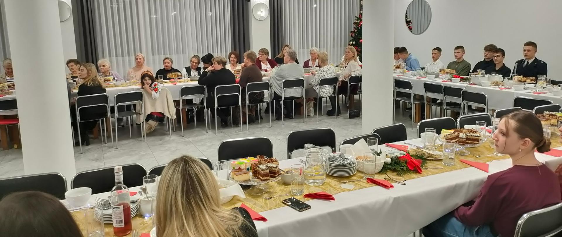Sala świetlicowa z długimi stołami ustawionymi w podkowę, przy których siedzą uczestnicy spotkania wigilijnego. Na stołach znajdują się białe obrusy, świąteczne dekoracje, ciasta, napoje i zastawa stołowa. W tle widoczna jest choinka oraz jasne, nowoczesne wnętrze z dużymi oknami i kolumnami. Uczestnicy rozmawiają i wspólnie spędzają czas przy świątecznym poczęstunku.