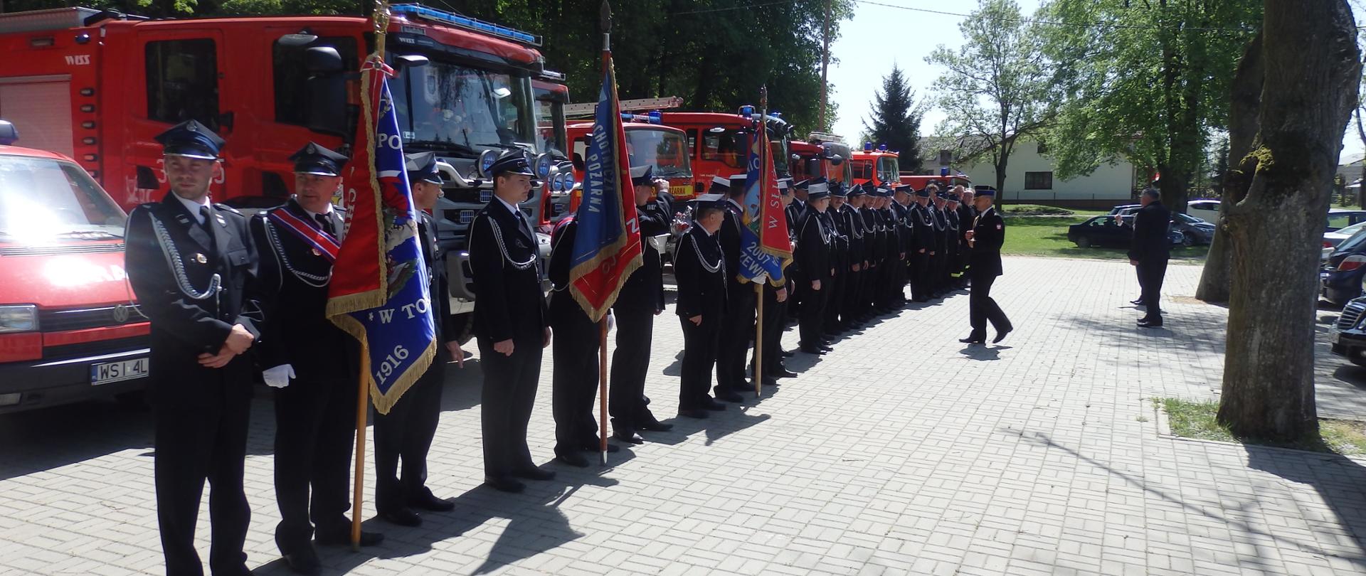 na tle samochodów strażackich grupa osób w mundurach strażaków, trzy osoby trzymają sztandary 
