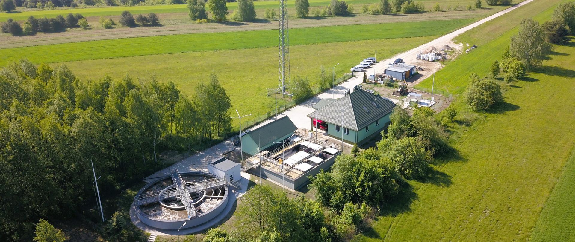 Widok z lotu ptaka na oczyszczalnie ścieków, duży okrągły reaktor, obok drugi mniejszy w kształcie prostokąta oraz dwa zielone budynki, wokół drzewa i pola.