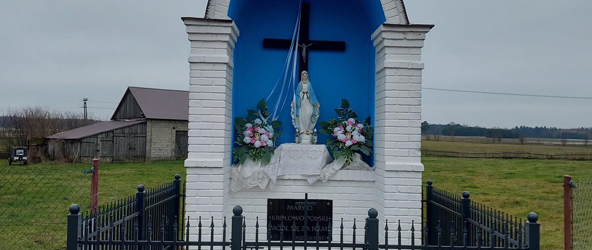 Zdjęcie przedstawia przydrożną murowaną kapliczkę znajdującą się przy ul. Łąkowej w Rudce. W kapliczce znajduje się figura Matki Boskiej. Kapliczka jest ogrodzona metalowym ogrodzeniem