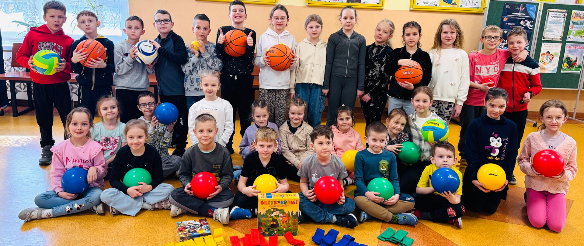 Grupa uczniów prezentująca nagrody zdobyte w ramach akcji "Szkoły pełne Talentów" (zestaw piłek, gry planszowe, szarfy)