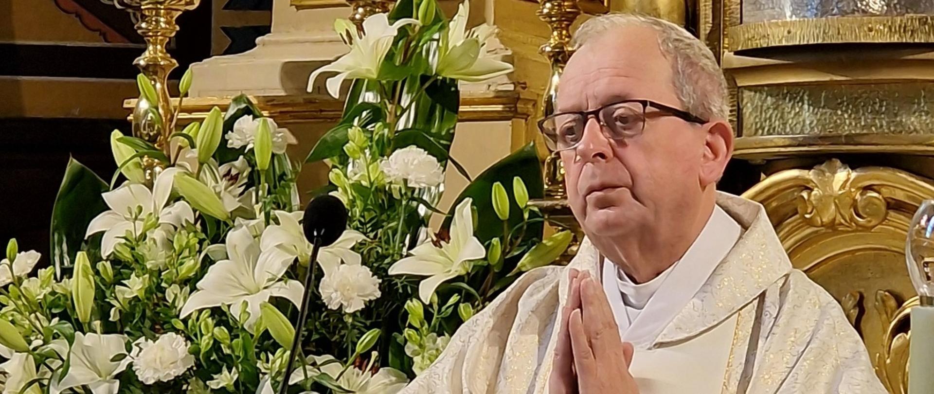 Proboszcz Wiesław Cygan w ornacie w kolorze ecru stoi zamyślony ze złożonymi do modlitwy rękami, przed nim mikrofon, za nim fragment ołtarza udekorowanego białymi liliami