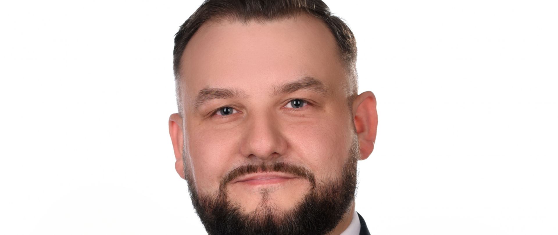 Mężczyzna włosy zaczesane do tyłu, kwadratowa twarz z ciemną brodą i wąsami, jasne łagodne oczy, lekki uśmiech. Ubrany w granatowy garnitur z jaśniejszą niebieską kratką do tego biała koszula i krawat