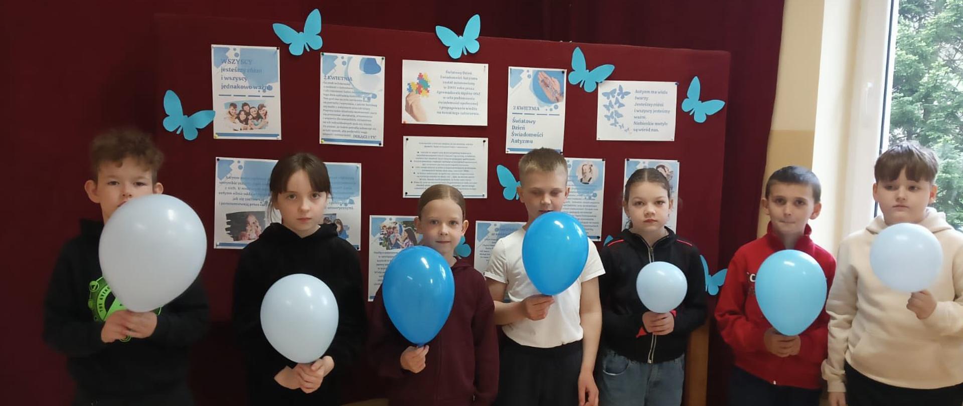 Uczniowie trzymają w ręku niebieskie balony, za nimi gazetka z informacjami na temat autyzmu