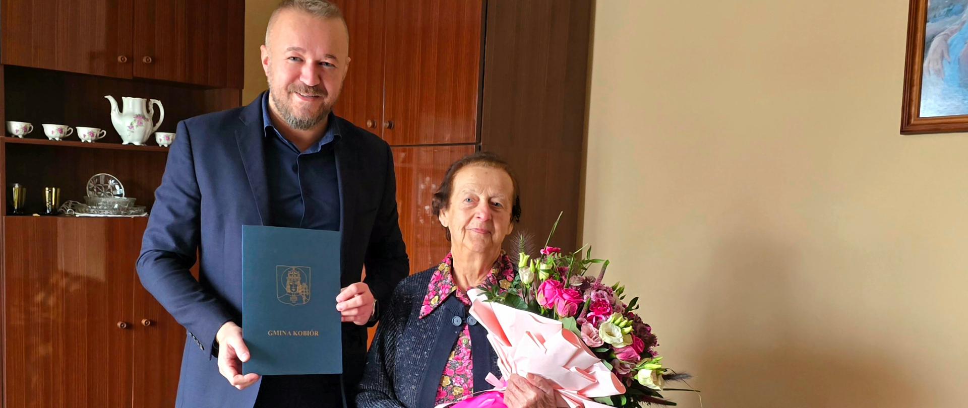Mężczyzna z kobietą trzymającą bukiet kwiatów pozują do zdjęcia na tle meblościanki.