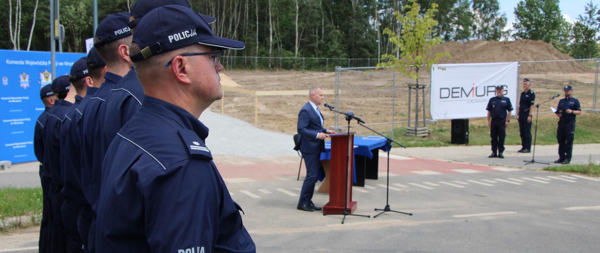 Mężczyźni ubrani w mundury stoją na baczność, w tle przemawia mężczyzna w garniturze. 