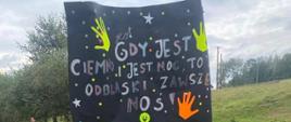 Uczniowie w kamizelkach odblaskowych idą poboczem, dwóch uczniów na przodzie niesie plakat promujący odblaski