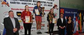 Trzy zawodniczki na podium po wręczeniu nagród w jednej z kategorii wagowych. Każda z nich ma zawieszony na szyi medal, a w rękach trzyma dyplom i puchar. Po lewej stronie podium stoi pan Jerzy Nieć, natomiast po prawej stronie zaraz przy trzecim stopniu podium stoi Przewodniczący Rady Powiatu w Kraśniku Jacek Dubiel.