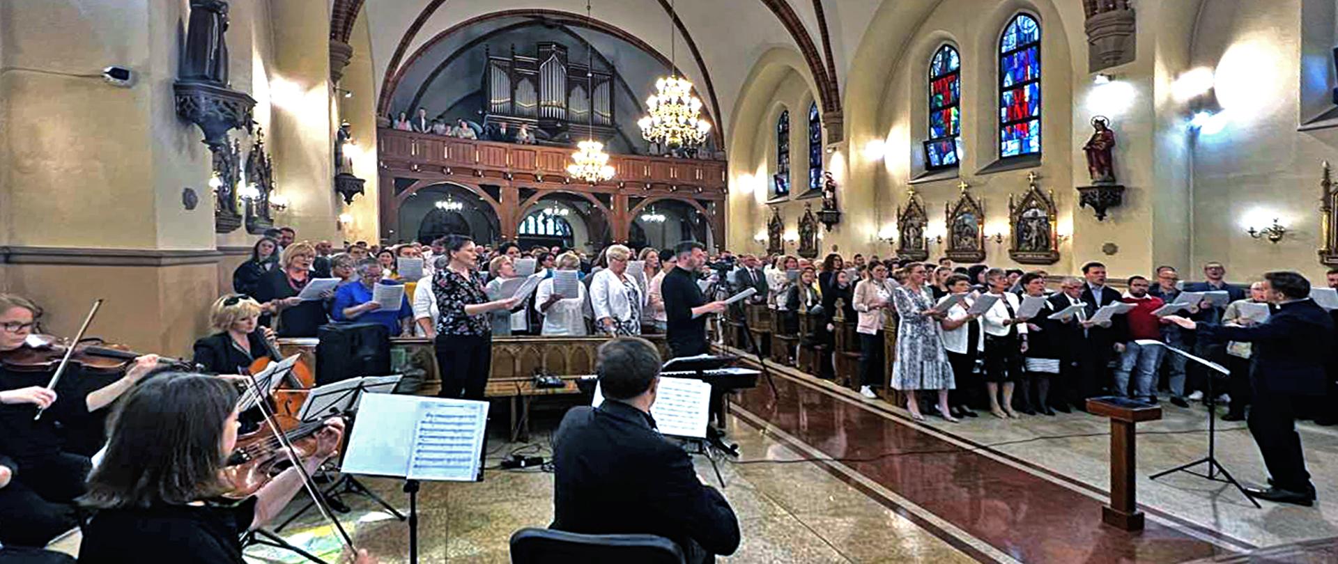 Na pierwszym planie grupa muzyków grająca na instrumentach smyczkowych, dalej grupa chórzystów w ławkach kościelnych w tle jasny wystrój kościoła z figurami świętych i stacjami drogi krzyżowej oraz zabytkowe organy.