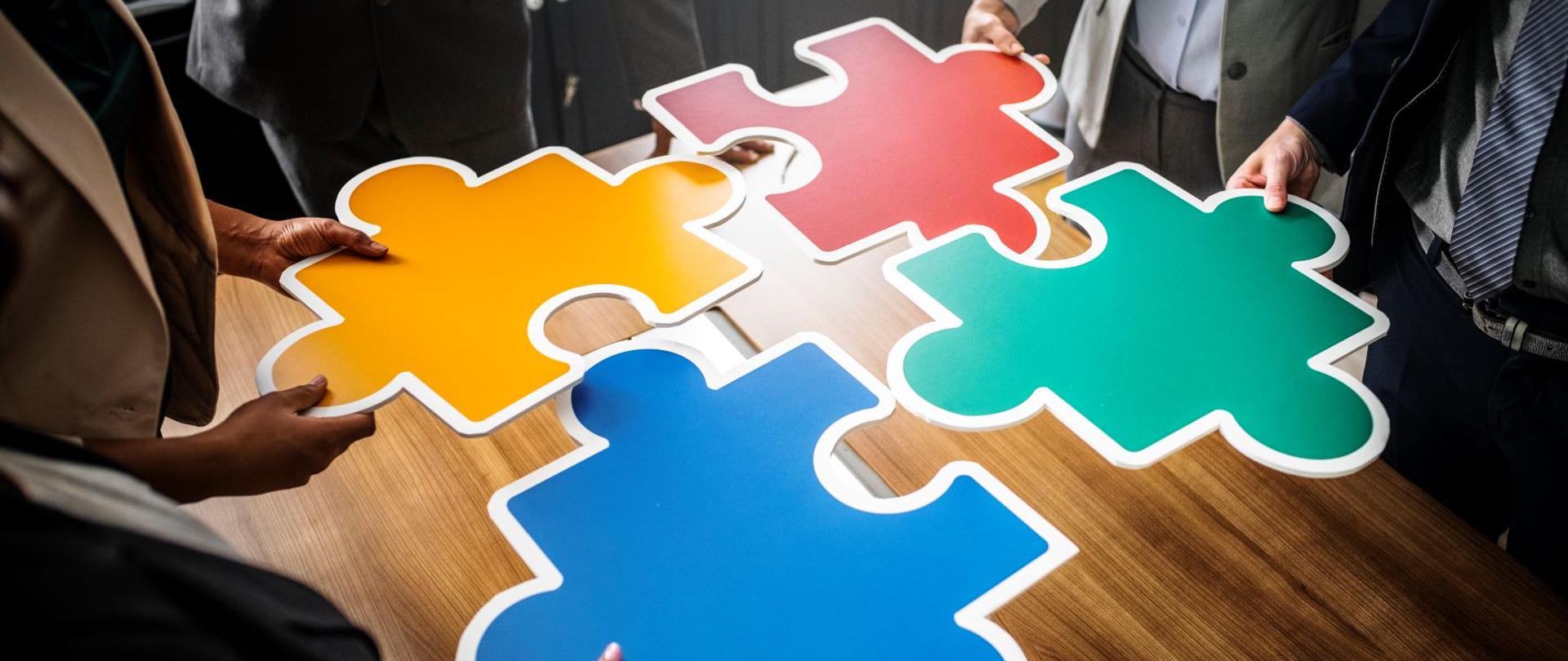 Ludzie trzymają cztery puzzle w kolorach niebieskim, żółtym, czerwonym i zielonym