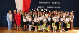 Zdjęcie grupowe uczestników i organizatorów wydarzenia na tle ścianki z biało -czerwoną flagą i nazwą konkursu.