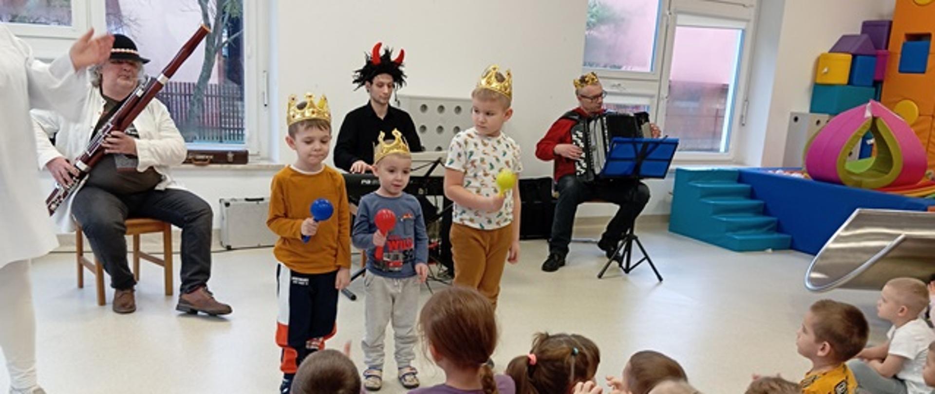 Zdjęcie prezentuje dzieci biorące udział w audycji muzycznej oraz teatrzyku. Przed grupą siedzących dzieci występują aktorzy