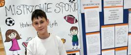 Uczeń w stroju sportowym trzyma w ręku piłkę nożną, za nim ścianka moderacyjna, na której przypięte zostały wykonane przez uczniów plakaty o sportowcach 