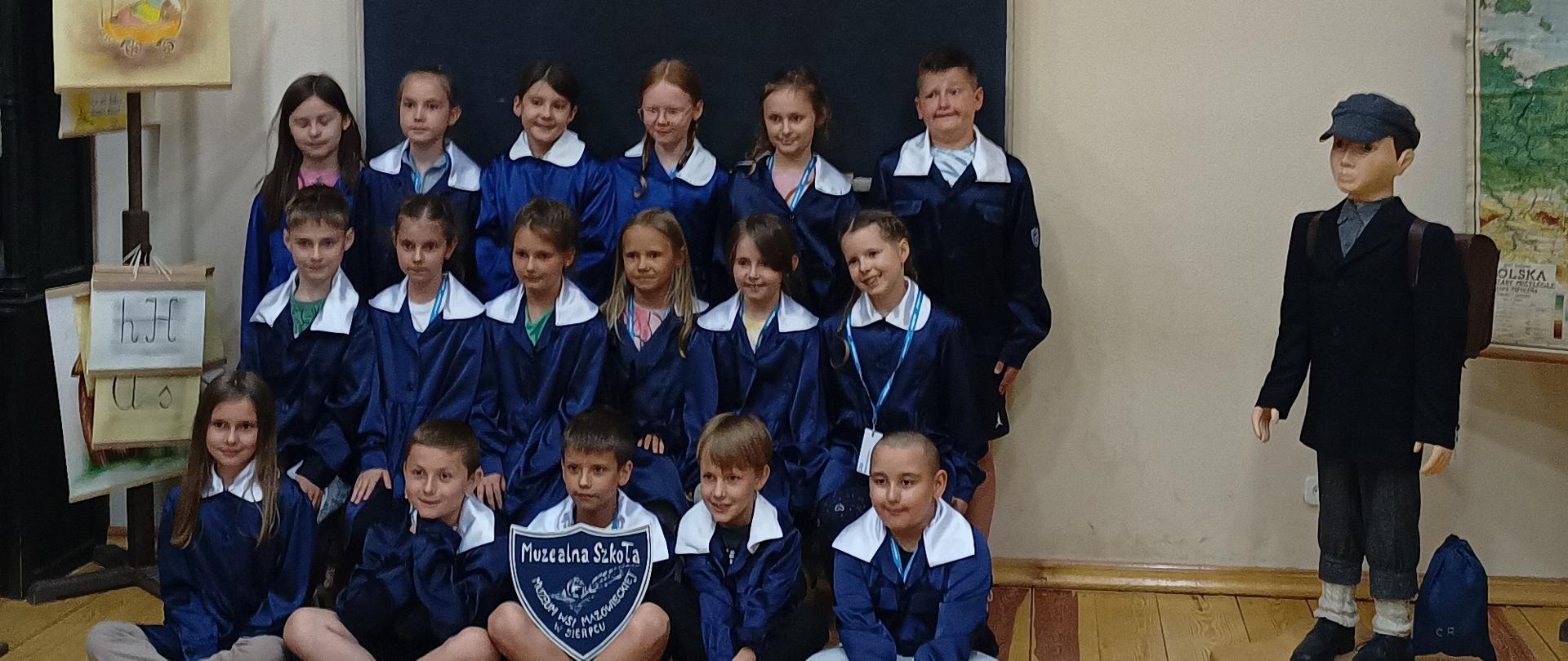 Grupa dzieci ubrana w granatowe mundurki z białymi kołnierzykami ustawiona w 3 rzędy. Pierwszy rząd siedzi na podłodze, pozostałe z tyłu stoją. Dziecko siedzące z przodu na środku trzyma tabliczkę z napisem Muzealna szkoła